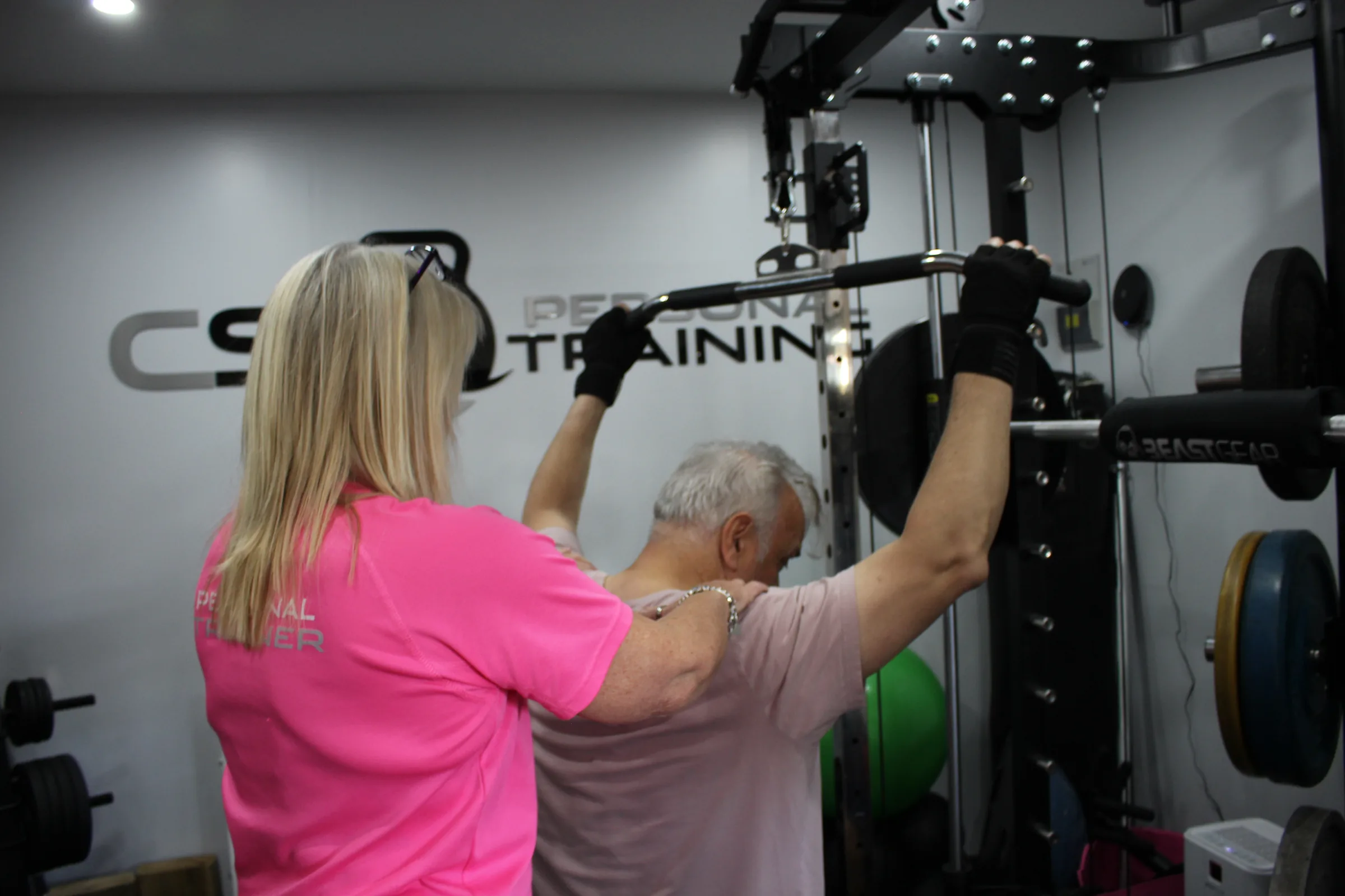 Carol with a hand on a client's shoulder blade as he pulls a cable bar down in the studio.