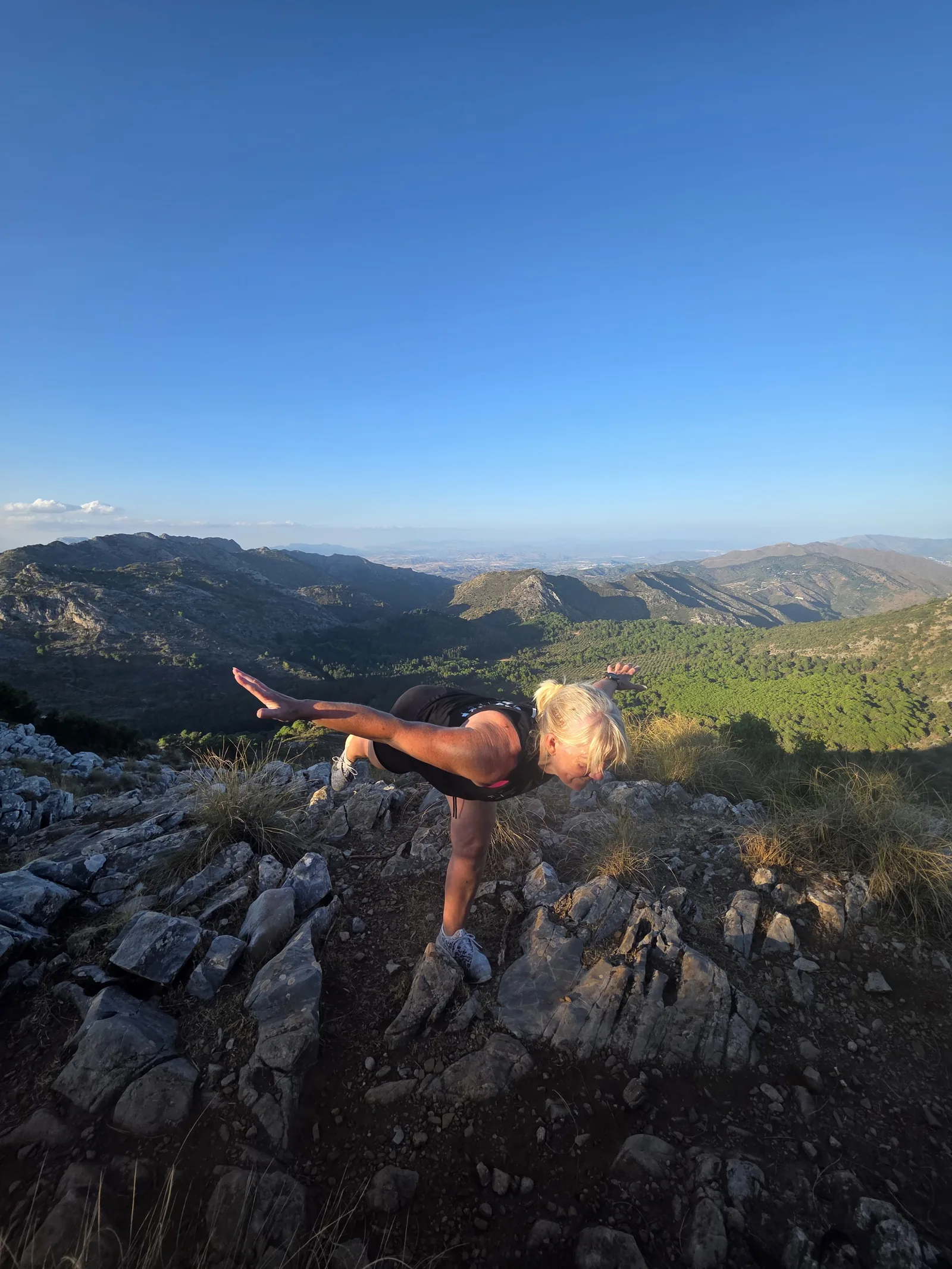 Carol holding a single leg balance on a rocky ridge, mountains behind her.